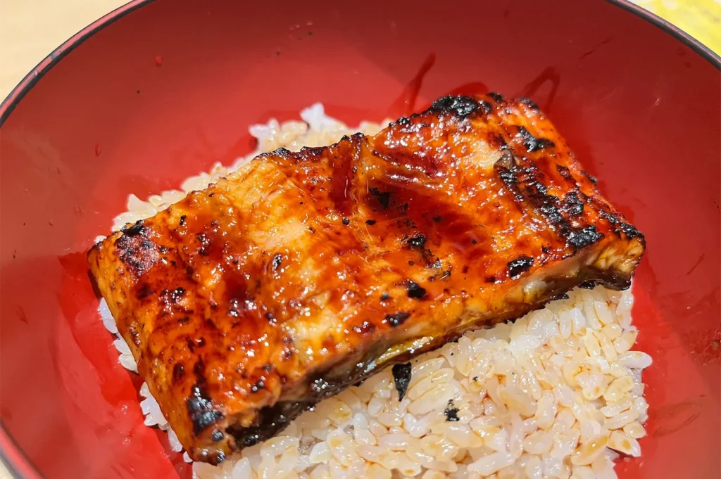 Close-up shot of a perfectly grilled, caramelized Unagi (freshwater eel) glazed with sweet soy tare sauce, served over a bed of steamed white rice in a traditional red bowl.