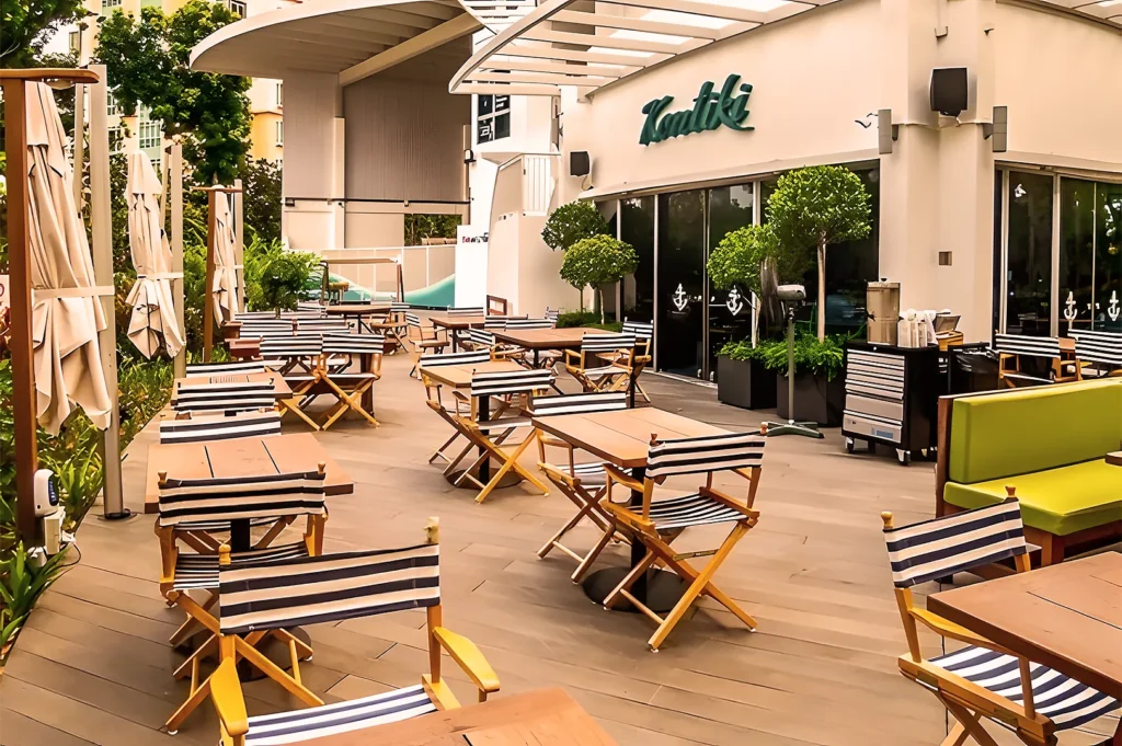 Outdoor patio of a restaurant with wooden tables, striped chairs, and lush greenery. A relaxed, welcoming ambiance under a modern, airy roof.