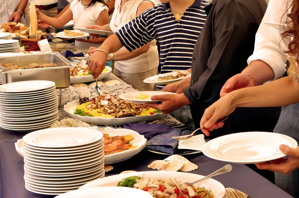 People serve themselves at a buffet, holding white plates and choosing from an array of colorful dishes, creating a warm, communal dining atmosphere.