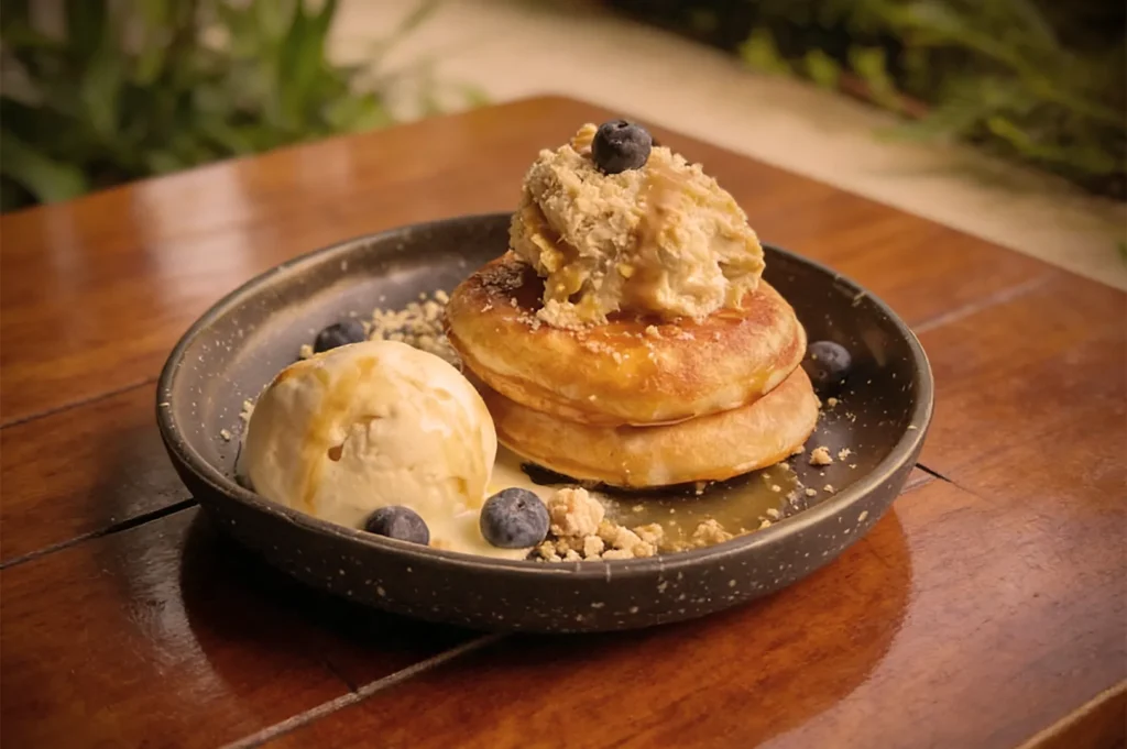 Fluffy pancakes topped with ice cream, granola, and blueberries on a dark plate. Next to them, a scoop of ice cream, creating a cozy, indulgent feel.