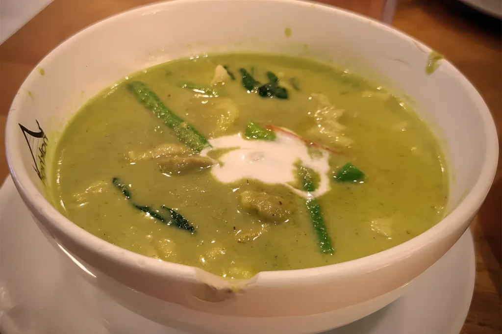 A rich and creamy bowl of Nakhon Kitchen’s Thai Green Curry with chicken, garnished with coconut milk swirl and fresh Thai basil leaves.