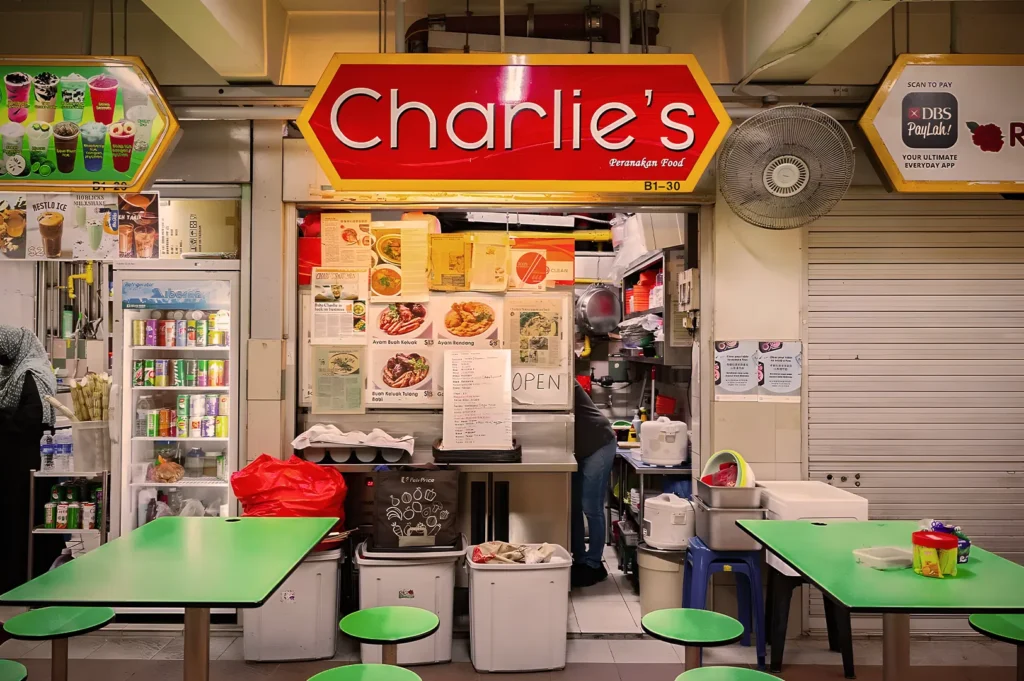 A vibrant food stall named "Charlie's" with a red sign, displaying menu pictures. Green tables and stools invite customers in a bustling market setting.