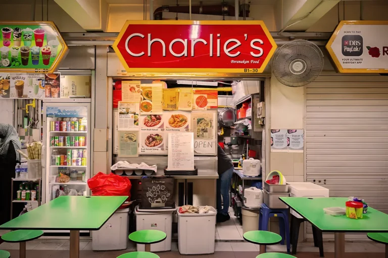 A vibrant food stall named "Charlie's" with a red sign, displaying menu pictures. Green tables and stools invite customers in a bustling market setting.