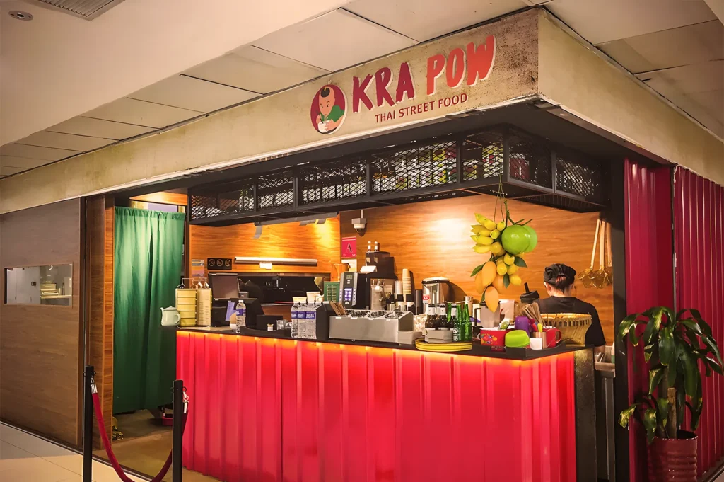 Kra Pow Thai Street Food stall with a vibrant red counter, hanging fruit decorations, and an open kitchen preparing authentic spicy dishes.