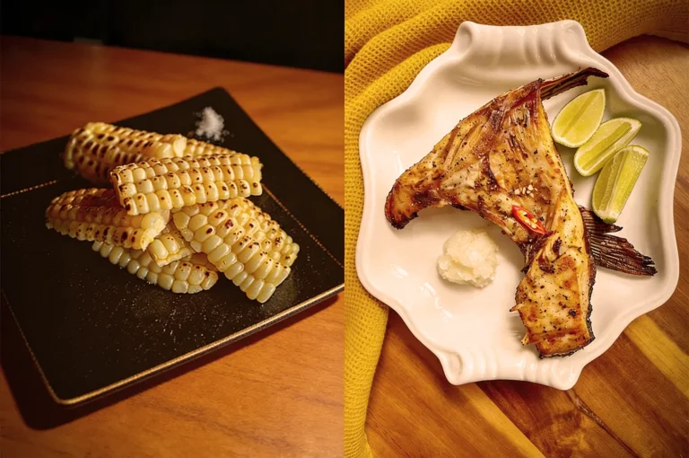 Grilled corn pieces stacked on a black plate beside grilled fish with lime wedges on a white plate, atop a wooden table with a yellow cloth.