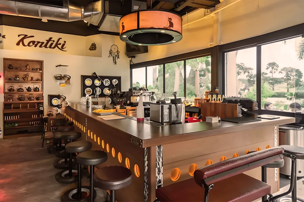 Cozy cafe interior with a central espresso bar featuring stools, warm lighting, and retro decor. Large windows reveal a greenery-filled view outside.