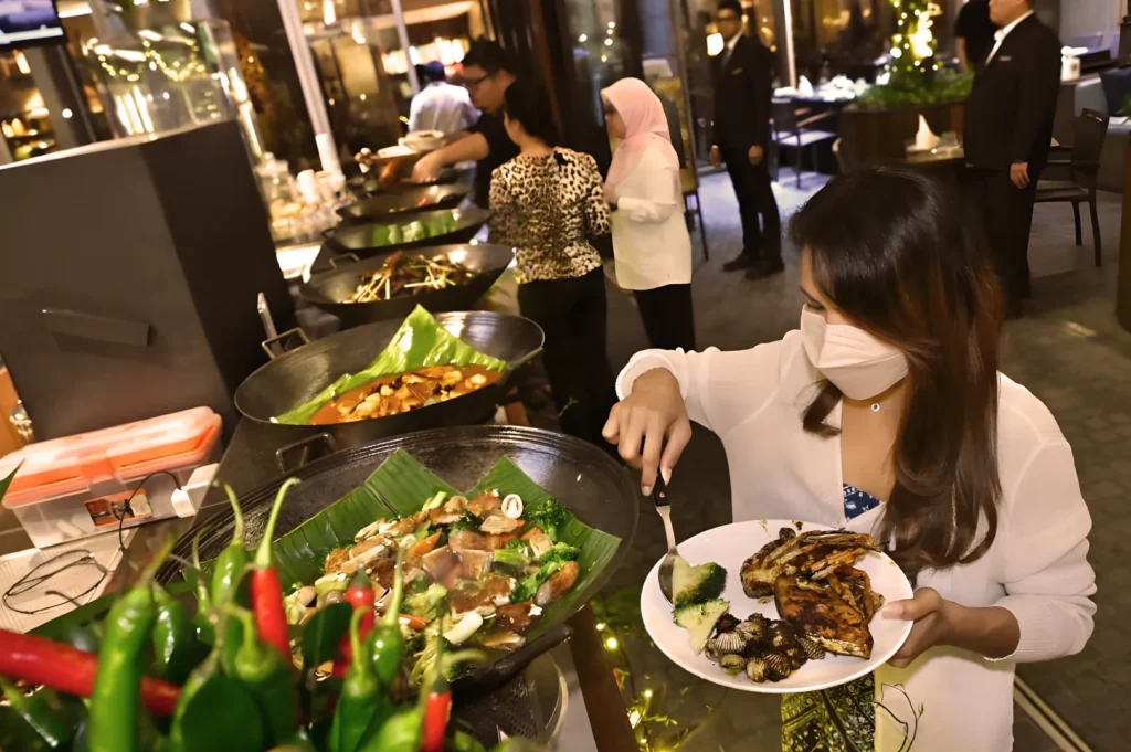 A woman in a white shirt and mask serves herself grilled food at a buffet with large dishes of various cuisines. The atmosphere is lively and elegant.