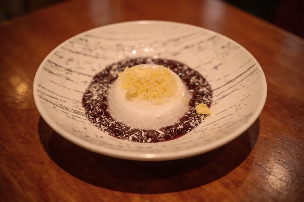 Creamy panna cotta topped with crumbles sits in a white dish, surrounded by a rich berry sauce. The wooden table adds a warm, rustic feel.