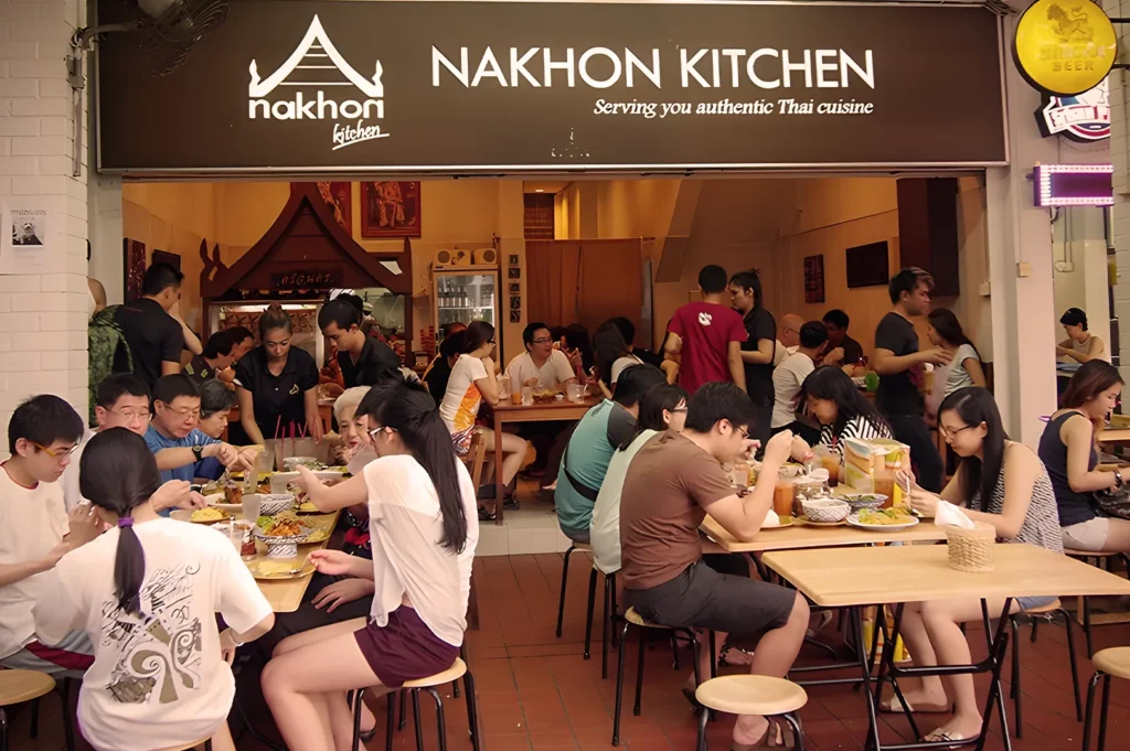Bustling evening crowd at Nakhon Kitchen, showing a popular open-concept Thai eatery filled with diners and a prominent "Serving you authentic Thai cuisine" sign.