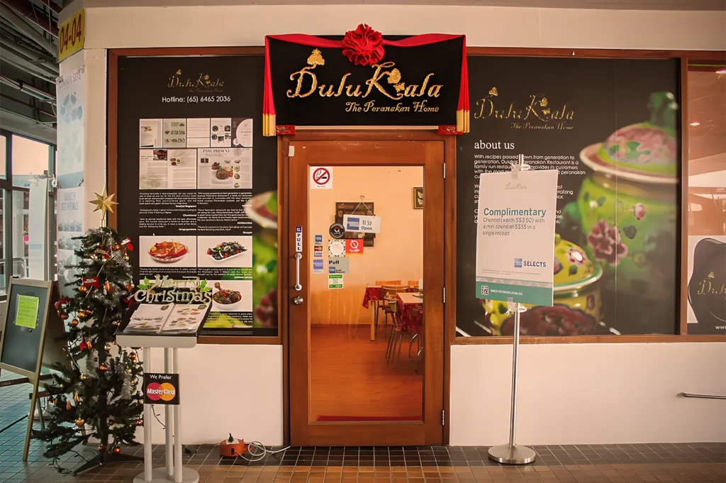 Shop entrance of "Dulukala The Peranakan Home" with Christmas decor; includes a decorated tree, glass door, and promotional signs, creating a warm, festive atmosphere.