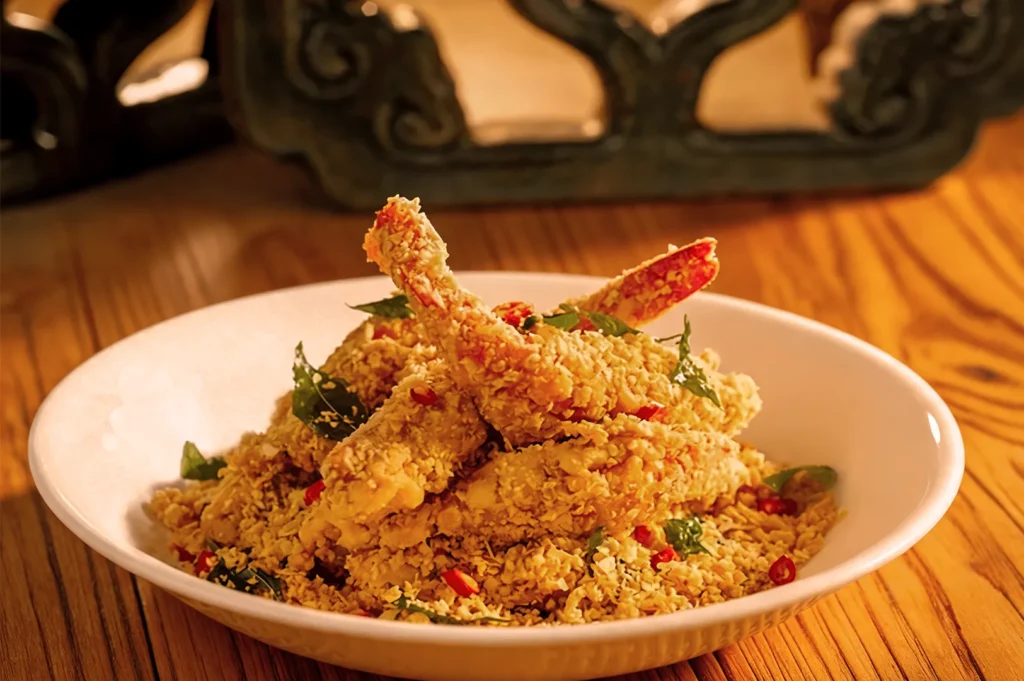 A bowl of golden, crispy crab legs garnished with red chili slices and leafy greens, set on a wooden table, conveying a warm, appetizing feel.