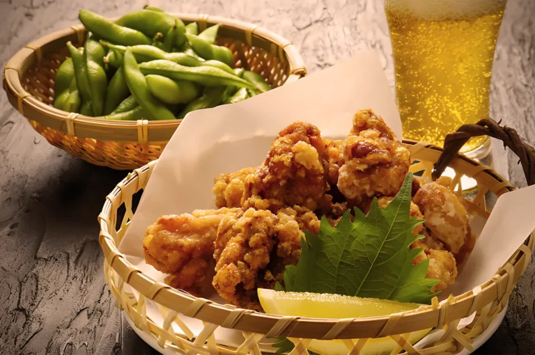 A basket of crispy fried chicken with a lemon wedge and leaf garnish sits on a table. Next to it are edamame beans and a glass of frothy beer.