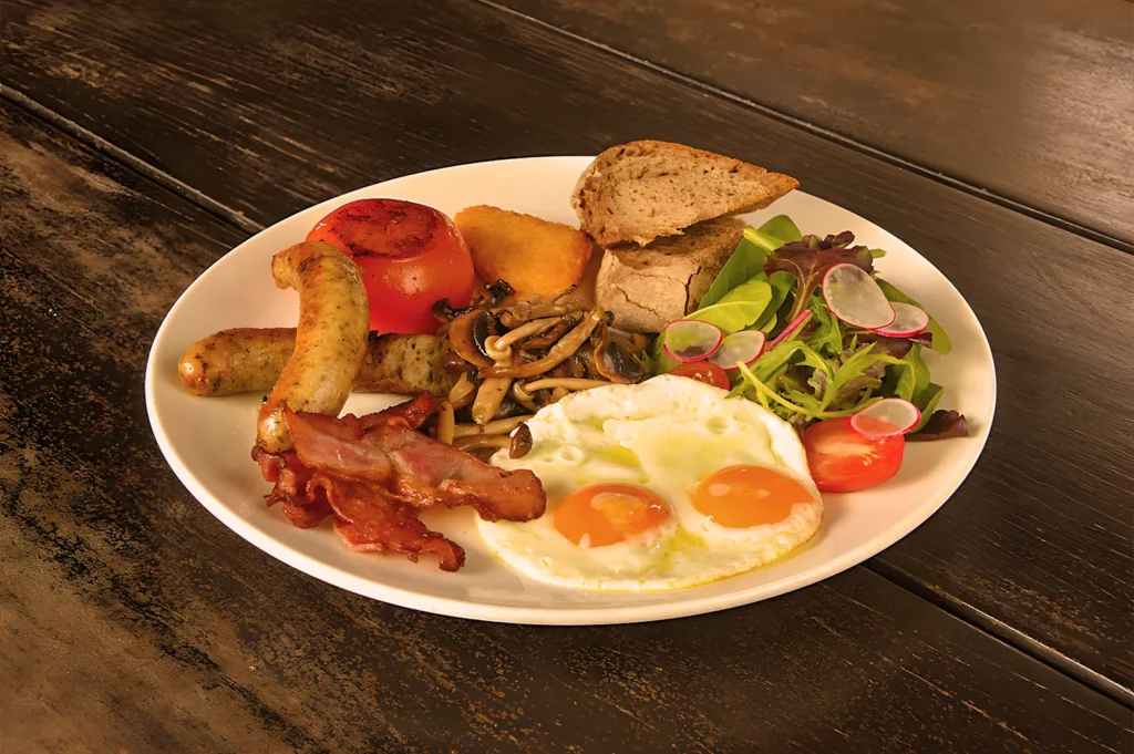 Plate of breakfast food on a wooden table with eggs, bacon, sausage, grilled tomato, mushrooms, toast, fresh greens, and radishes. Inviting and hearty.