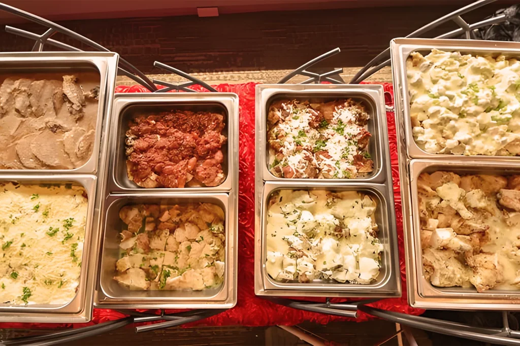 A buffet with trays of various foods including sliced meats, baked pasta, and creamy vegetable dishes, garnished with herbs on a red tablecloth.
