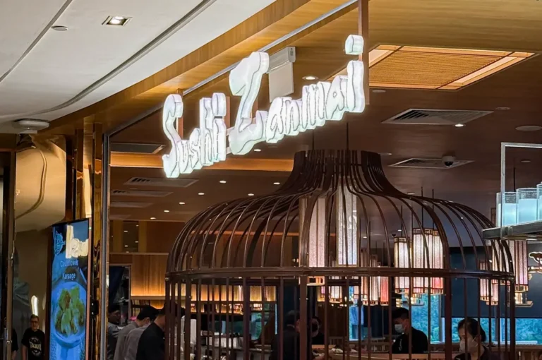 The glowing Sushi Zanmai neon sign above a unique birdcage-style wooden structure inside a modern Japanese dining area.
