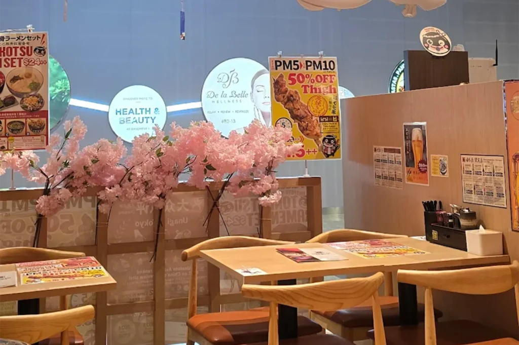 Indoor dining section of a Japanese restaurant featuring minimalist light wood tables, chairs, pink artificial cherry blossoms, and promotional flyers in the background.