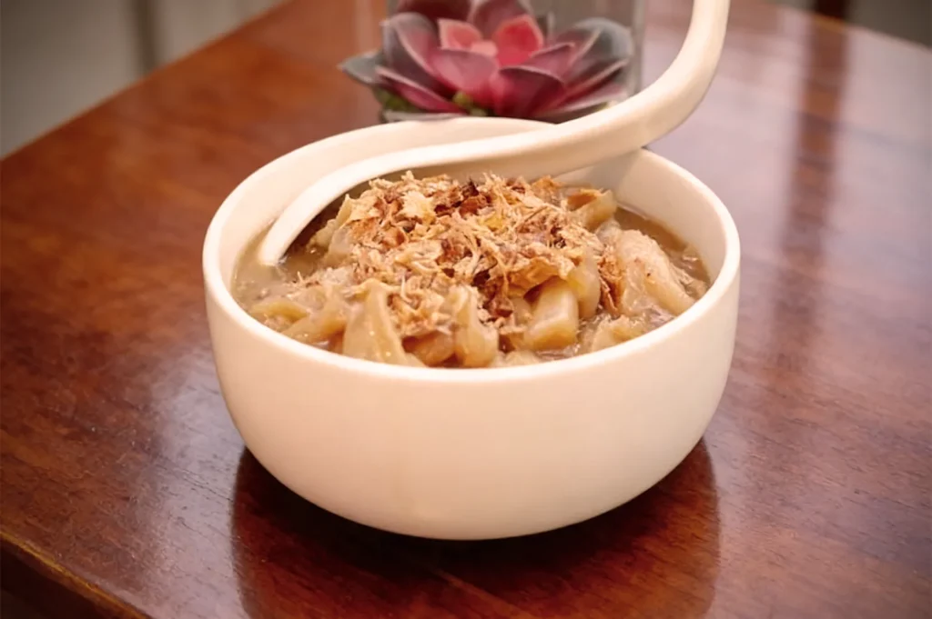 A white bowl on a wooden table filled with savory tripe soup, garnished with crispy fried onions. A ceramic spoon is resting in the bowl.