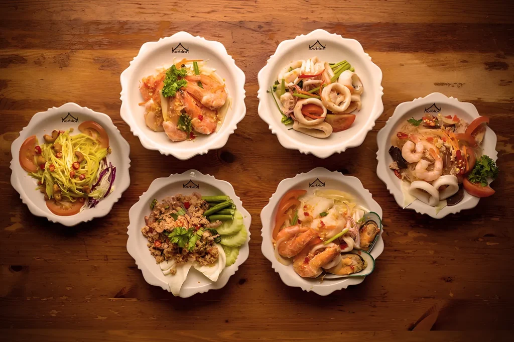 Overhead view of a Thai food spread from Nakhon Kitchen including Mango Salad, Larb Moo, Stir-fried Squid, and glass noodle seafood salad on a wooden table.