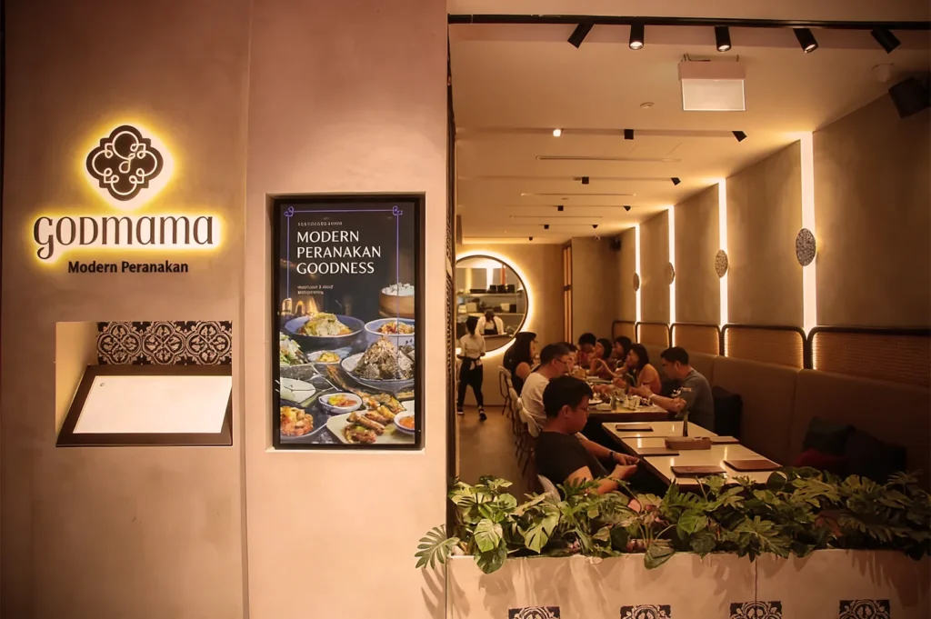 Cozy restaurant named Godmama, featuring "Modern Peranakan Goodness" poster, warm lighting, patrons dining, and lush green plants.