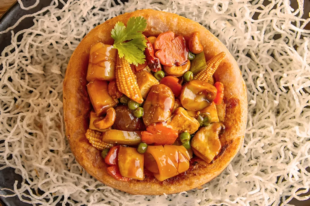 A savory dish featuring stir-fried vegetables, baby corn, carrots, and bell peppers in a crispy, edible bowl, garnished with a cilantro leaf. Surrounded by crispy noodles on a dark plate, creating an appetizing, colorful presentation.