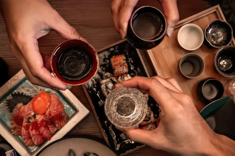 Three hands hold sake cups over a wooden table with sushi and sashimi dishes. The scene conveys a celebratory and communal atmosphere.