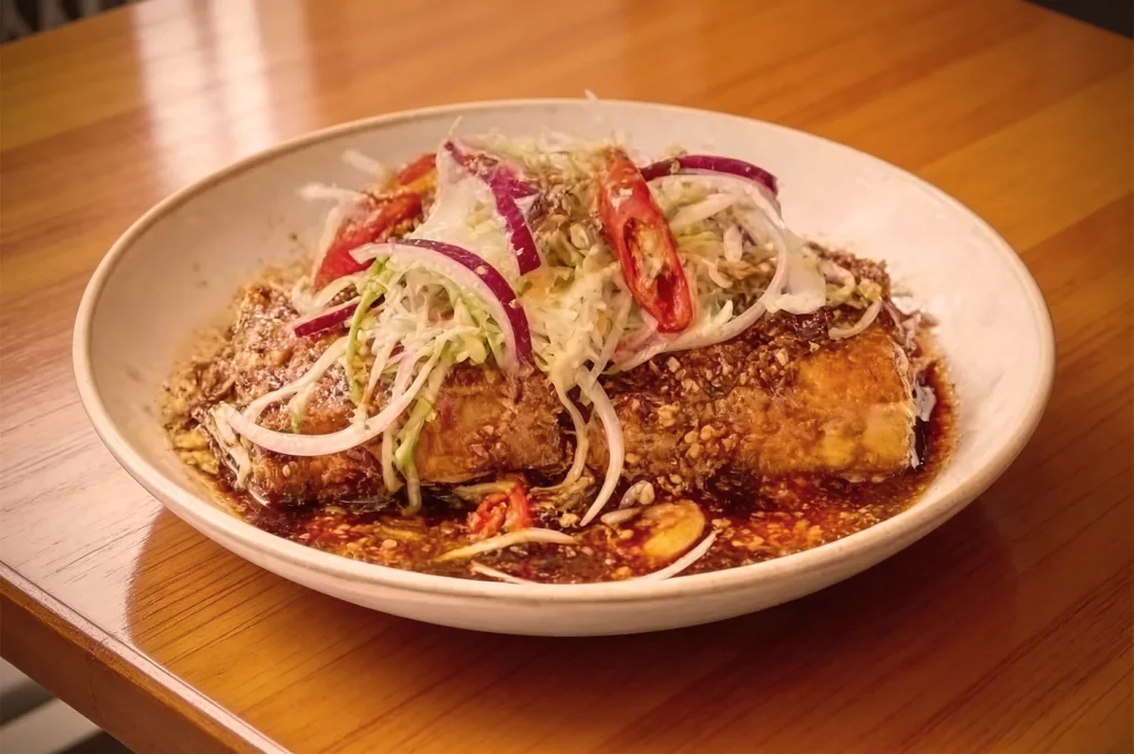 A white plate holds a crispy fried dish topped with sliced red onions, chili peppers, and shredded cabbage, drizzled with a savory sauce.