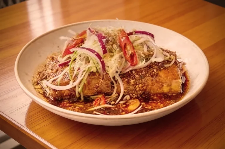 A white plate holds a crispy fried dish topped with sliced red onions, chili peppers, and shredded cabbage, drizzled with a savory sauce.