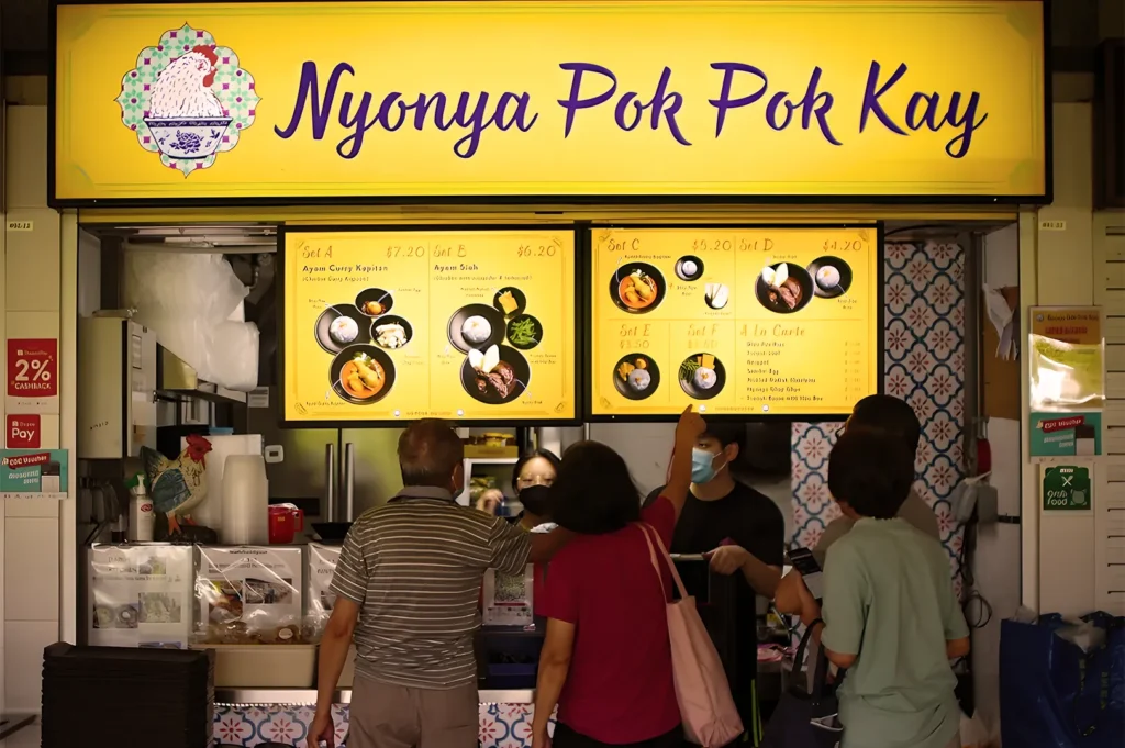 A group of people stands in front of a vibrant yellow food stall with the sign "Nyonya Pok Pok Kay." The menu displays colorful dish images.