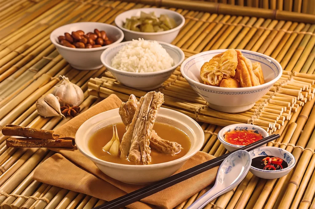 A traditional Chinese meal on bamboo mat, featuring a bowl of herbal soup with meat, accompanied by rice, nuts, pickles, fried dough, and sauces.