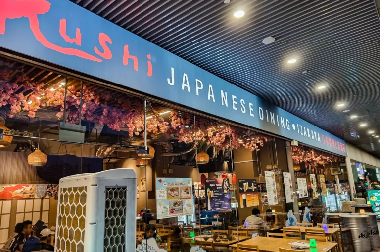 The blue facade of Kushi Japanese Dining and Izakaya featuring cherry blossom decorations, outdoor seating area, and posters for food specials.