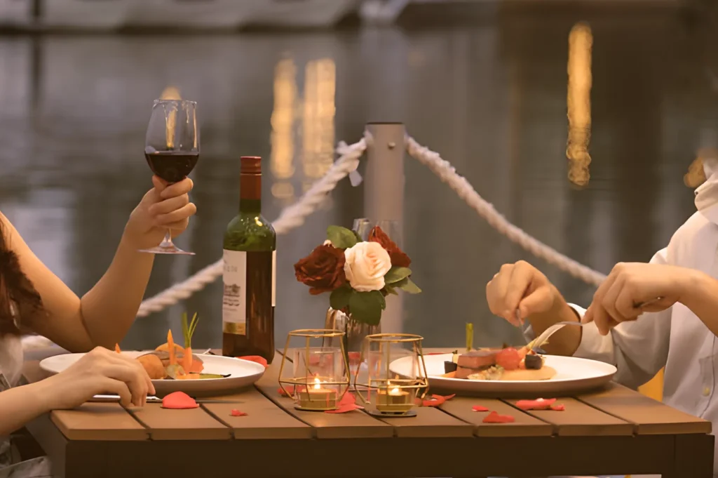A couple enjoys a romantic dinner by a lakeside, with red wine, candles, and rose petals on a wooden table, creating a warm and intimate atmosphere.