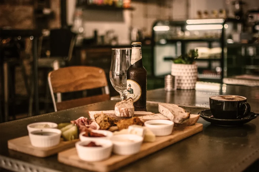 Cozy café setting with a wooden board of cheeses, bread, and meats, a beer bottle and glass, plus a cup of coffee on a metal table. Warm, rustic ambiance.