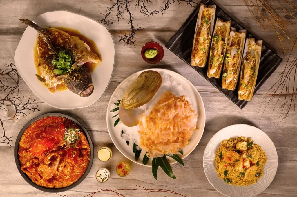 A variety of seafood dishes on a wooden table. Includes a grilled fish, clams, shrimp pasta, and a red curry dish, garnished with herbs. Rustic and appetizing.