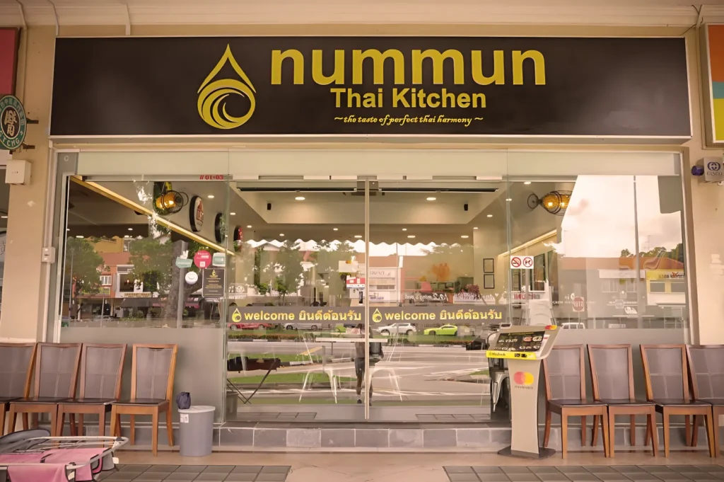 Modern exterior of Nummun Thai Kitchen with a prominent yellow logo, clean glass doors, and comfortable waiting chairs for diners.