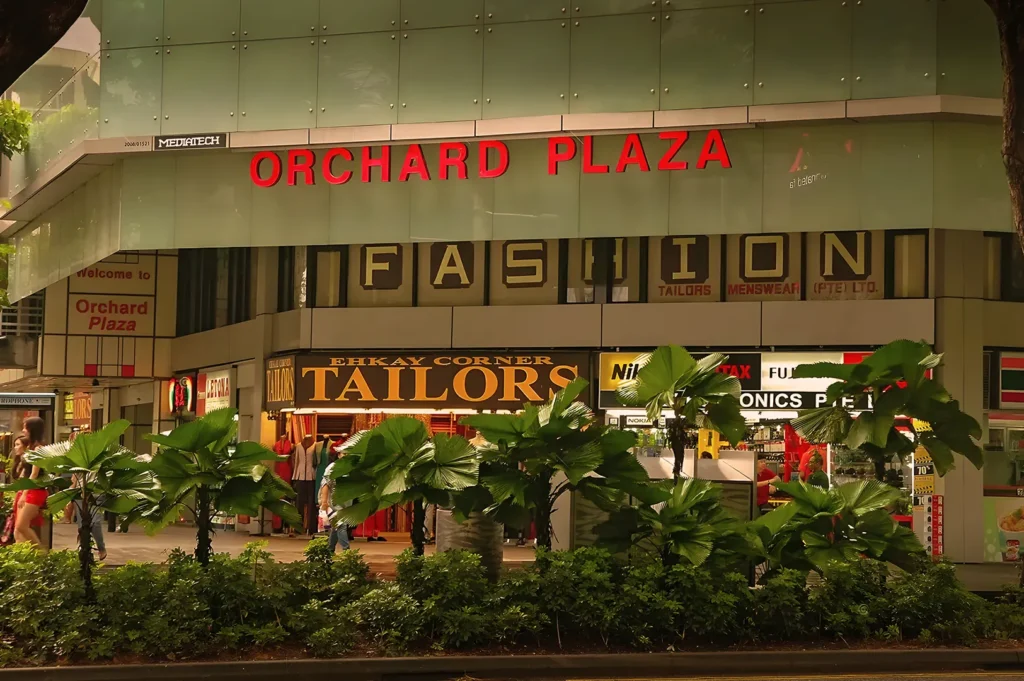 Exterior of Orchard Plaza, featuring bold red signage and various storefronts. Lush green plants line the sidewalk, adding a vibrant feel.