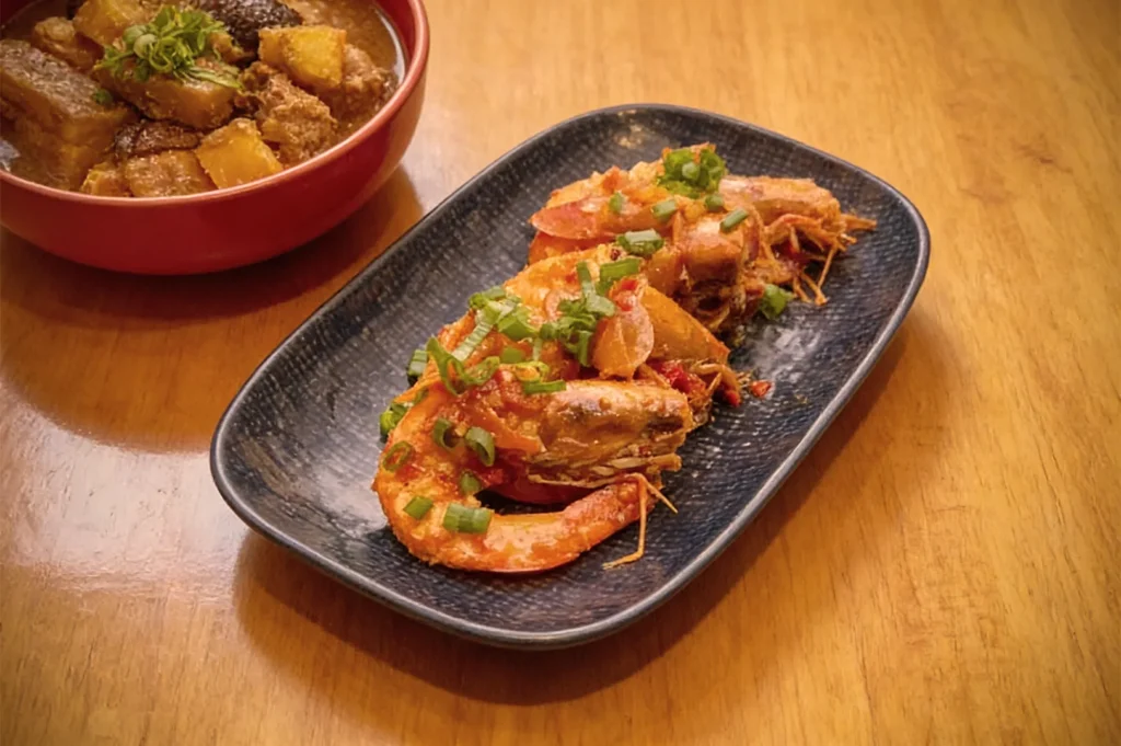 Stir-fried shrimp topped with green onions on a dark plate, next to a bowl of savory pork stew with potatoes, set on a wooden table.