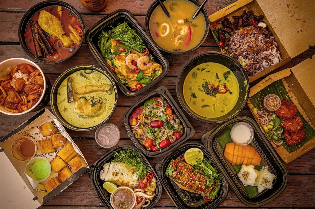 An overhead spread of diverse Thai takeaway dishes including Green Curry, Tom Yum Soup, Mango Sticky Rice, and grilled meats in eco-friendly containers.