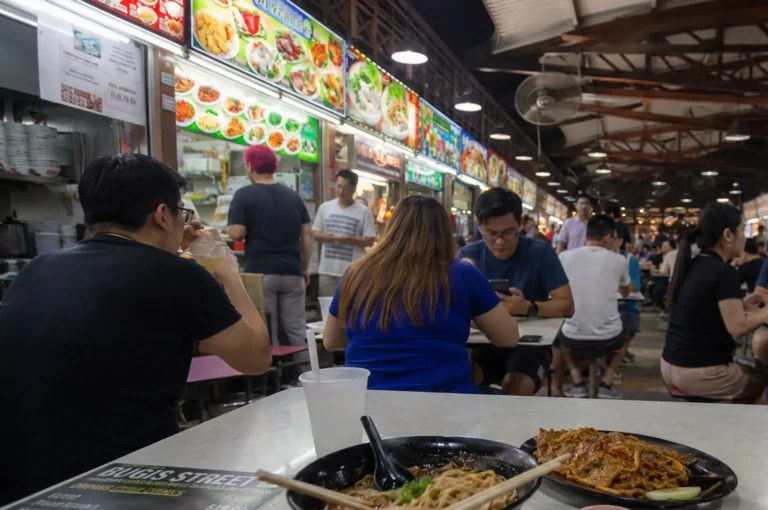 Busy hawker center scene at Bugis Street with diners eating at tables, illuminated food stall signs in the background, and plates of noodles in the foreground