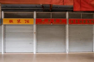 Closed storefronts with metal roller shutters and Chinese-English signs in an older shopping center