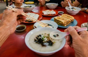 Family sharing bowls of porridge, chee cheong fun, and toast at a red hawker table.