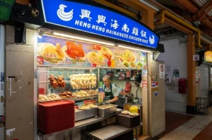 Heng Heng Hainanese Chicken Rice hawker stall with a blue sign, displaying hanging steamed and roasted chickens while a worker prepares orders