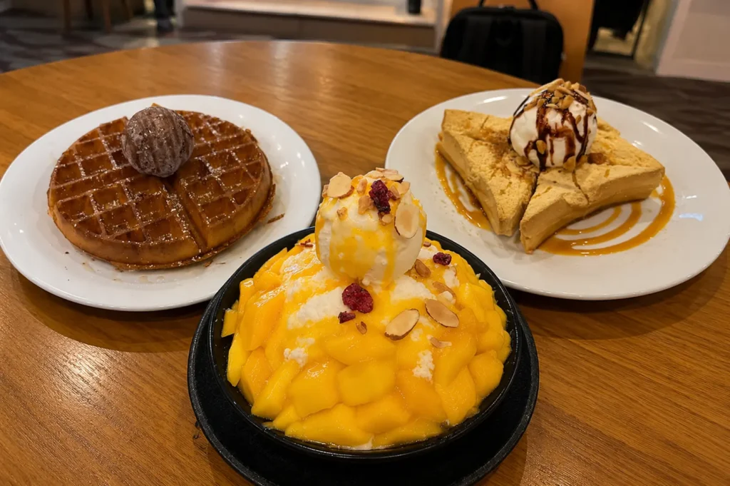 Table spread featuring a chocolate waffle, a mango bingsu topped with fresh fruit and ice cream, and a honey toast dessert
