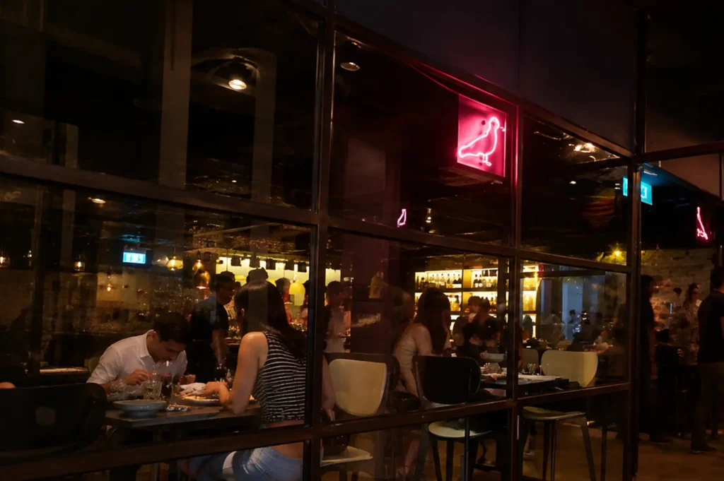 Exterior view of Neon Pigeon restaurant with lively evening diners behind glass