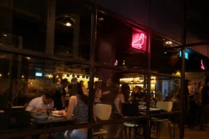 Exterior view of Neon Pigeon restaurant with lively evening diners behind glass