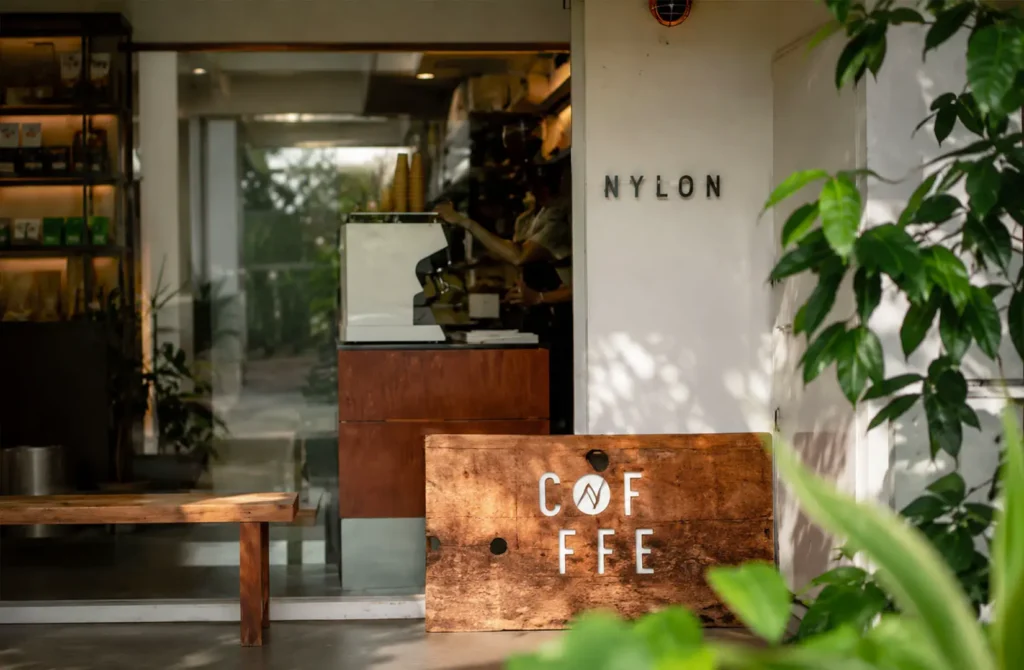 Entrance to Nylon Coffee featuring its minimalist storefront, wooden coffee sign, and lush greenery surrounding the café.