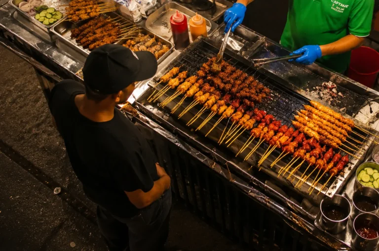 Street food vendor grilling and basting meat satay skewers over charcoal while a customer waits at a night stall
