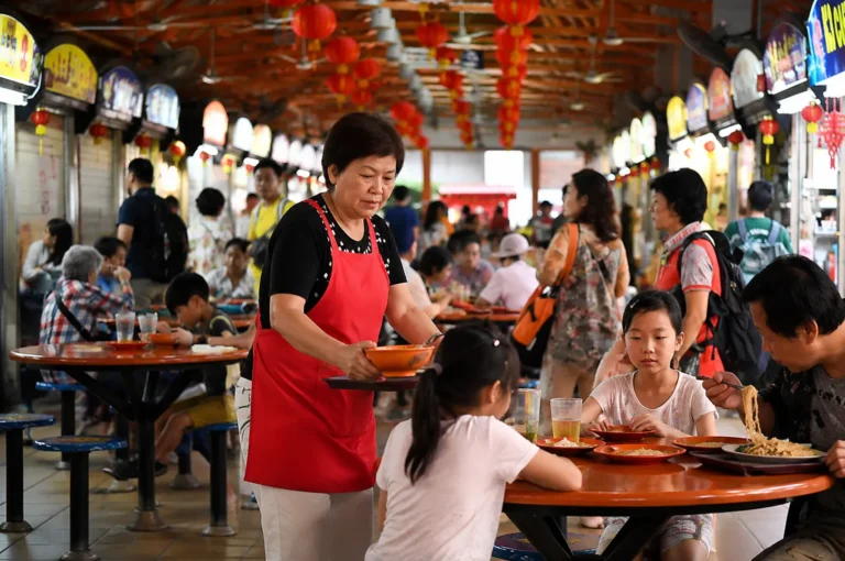 A busy hawker centre scene with a hawker in a red apron serving a family at a round table under hanging red lanterns