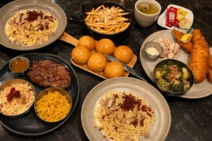 Western food spread featuring pasta carbonara, grilled steak, french fries, bread rolls, fish and chips, and side dishes