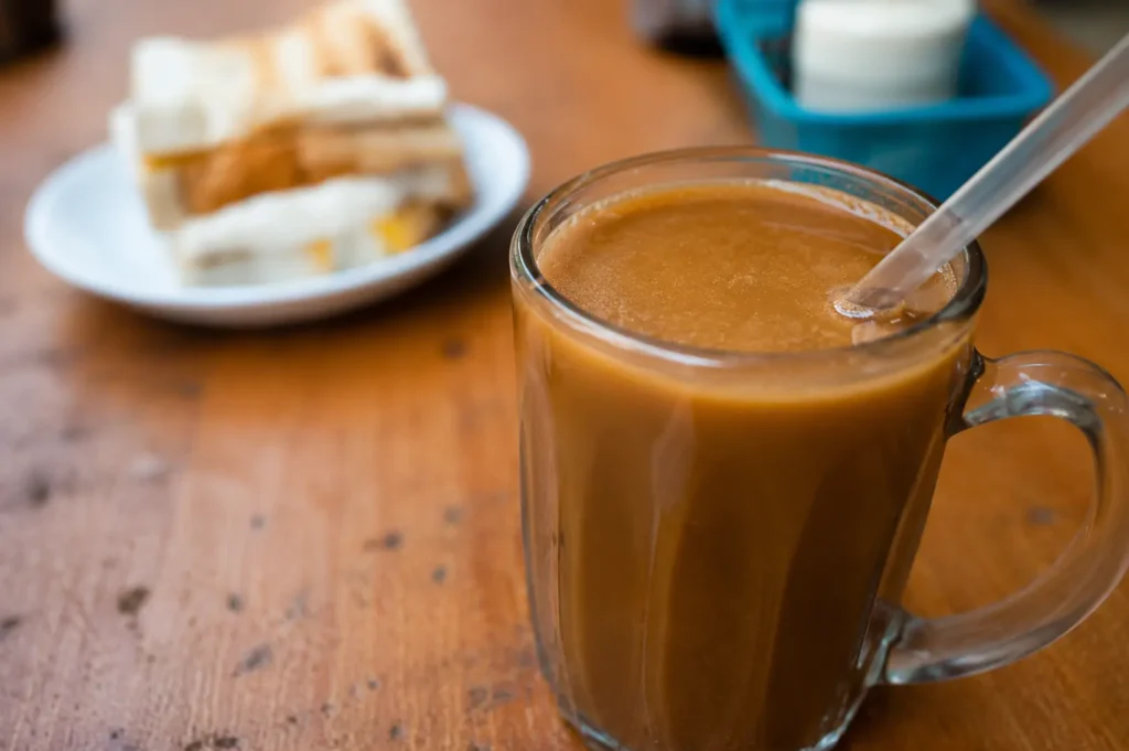 Glass mug of traditional kopi with kaya toast on wooden table at kopitiam