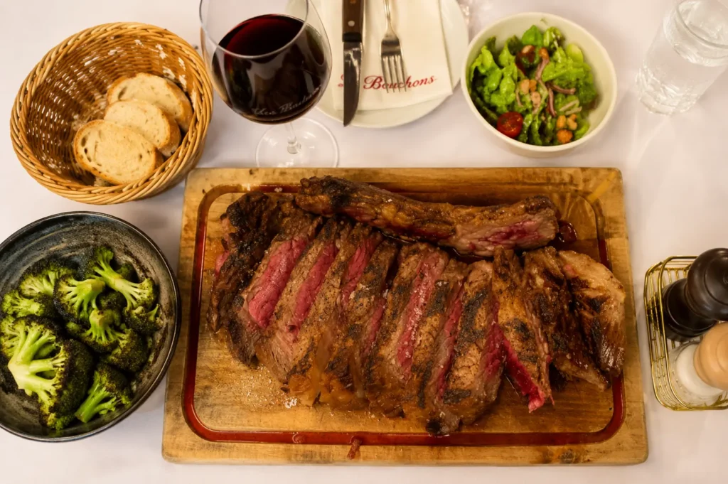 Overhead view of a sliced steak on a wooden board served with broccoli, bread, salad, and a glass of red wine
